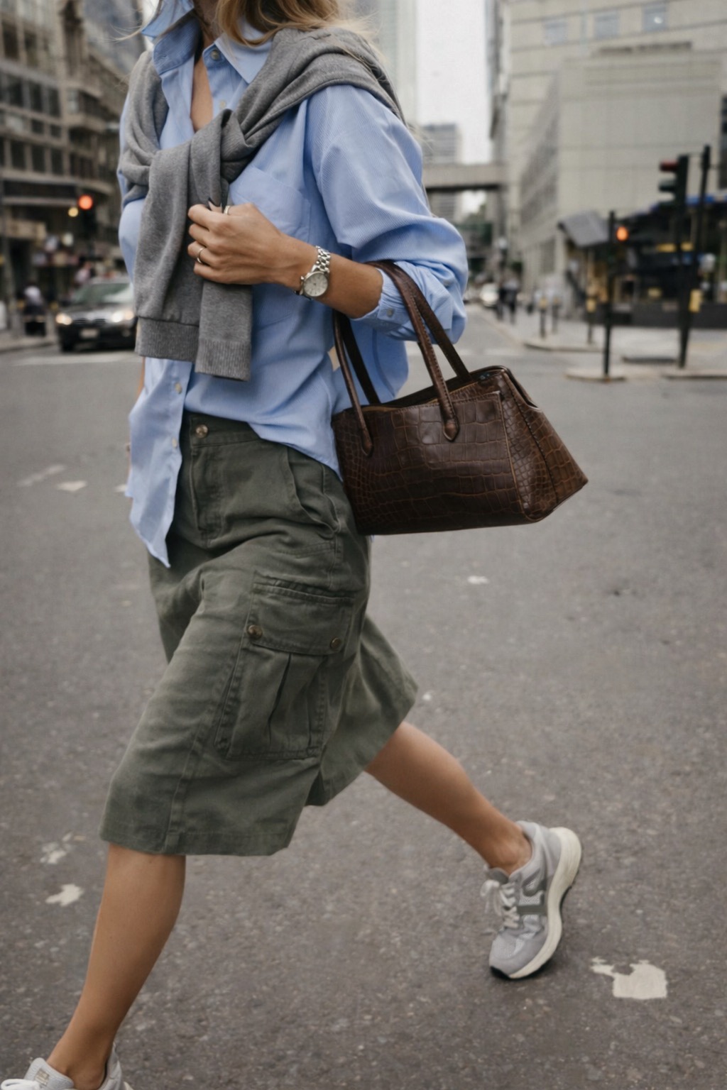 Blue oxford shirt, grey knit tied over the shoulders and olive cargo skirt, mid-stride in the city